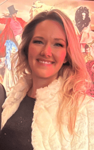 Portrait of a smiling woman with long blonde hair, wearing a white fur jacket, against a colorful background featuring circus-themed artwork, representing the team at Balance Process alongside Adam Lesner, mortgage loan officer.