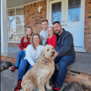 Family portrait on a front porch featuring a couple with three children and a golden doodle dog, showcasing a warm, friendly atmosphere.